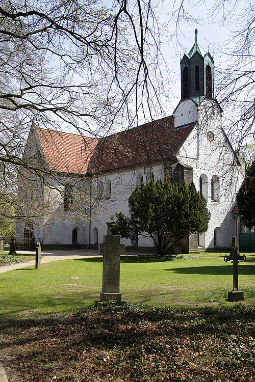 Die Seitenansicht einer romanischen Basilika mit heller Fassade und rotem Dach, im Vordergrund sind Grabsteine zu sehen.