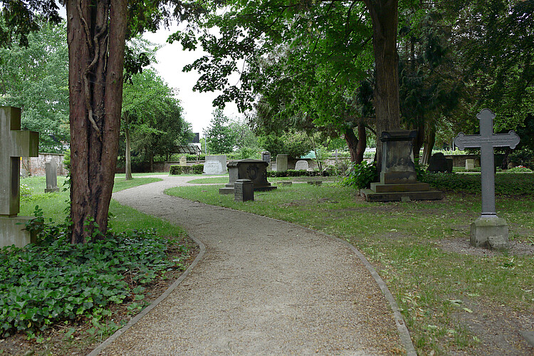 Ein geschotterter Weg führt in einem leichten Schwung über den Friedhof Marienwerder mit Grabsteinen und Grabkreuzen.