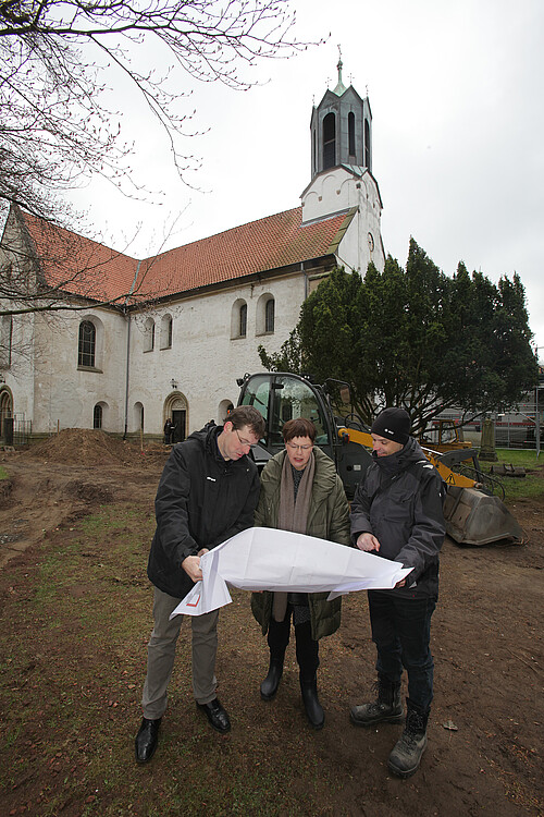 Drei Personen stehen mit einem großen Plan in den Händen vor der Kirche Marienwerder.