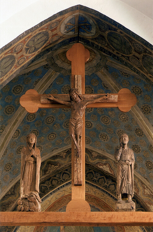 Ein hölzernes kreut mit einer Darstellung Jesu, an beiden Seiten sind weitere Figuren aus Holz dargestellt.