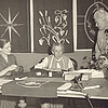 Drei Frauen sind um einen Tisch herum gruppiert, auf diesem liegen unterschiedliche Stickarbeiten.
