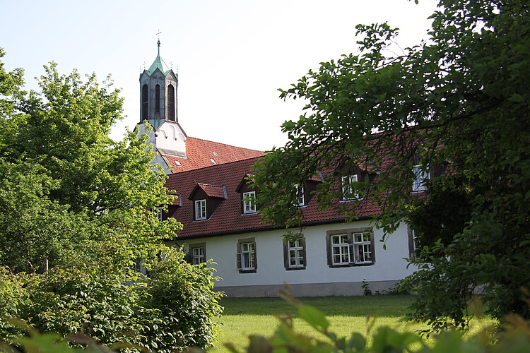 Seitenansicht einer hellen Gebäudefassade mit rotem Dach aus der Barockzeit. Im Vordergrund sind Bäume, im Hintergrund der Reiter einer Kirche zu sehen.