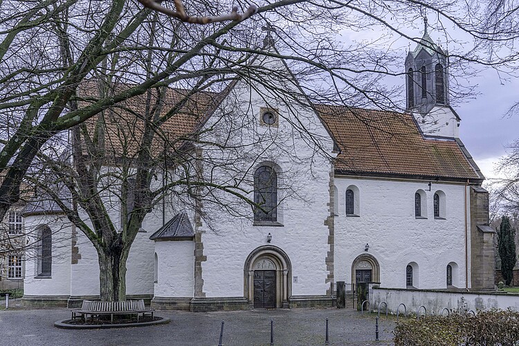 Die Seitenansicht einer romanischen Basilika mit heller Fassade und rotem Dach, im Vordergrund sind Grabsteine zu sehen.